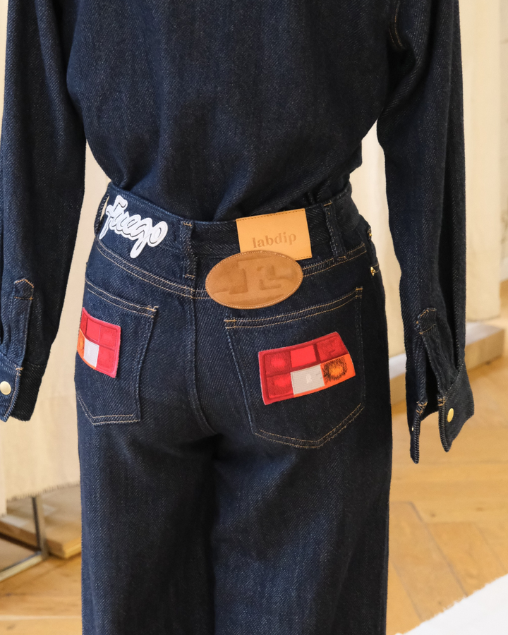 Jean en denim bleu foncé 100 % coton avec coupe droite et ajustée, poches arrière ornées de patchs rouges et détails de marque sur la taille.