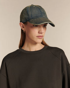 Femme portant une casquette en denim gris bleu et un vêtement noir, sur fond beige uni.