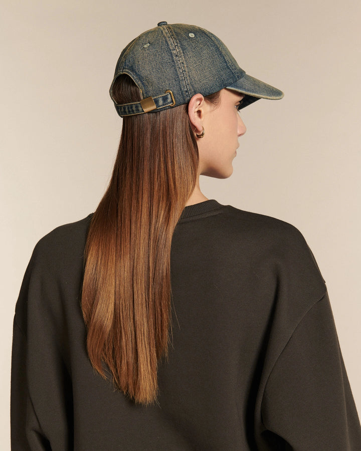Femme aux longs cheveux bruns portant une casquette en denim bleu délavé et un sweat noir uni sur fond beige.