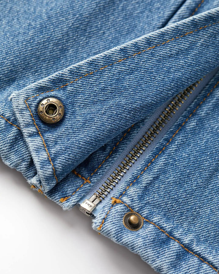Détail en gros plan d’un jean en denim bleu stone fade, 100 % coton, avec une fermeture éclair métallique et des boutons rivets apparents, surpiqûres orange contrastantes.
