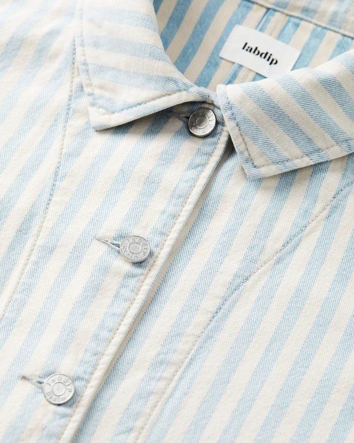 Détail d'une veste en denim léger rayé bleu ciel et écru, avec boutons métalliques gravés Labdip, tissu en coton structuré et coupe ajustée.