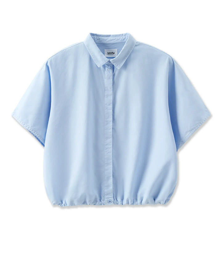 Chemise légère en popeline bleu clair, coupe ample et courte avec manches larges et col classique, tissu doux et fluide idéal pour l'été.