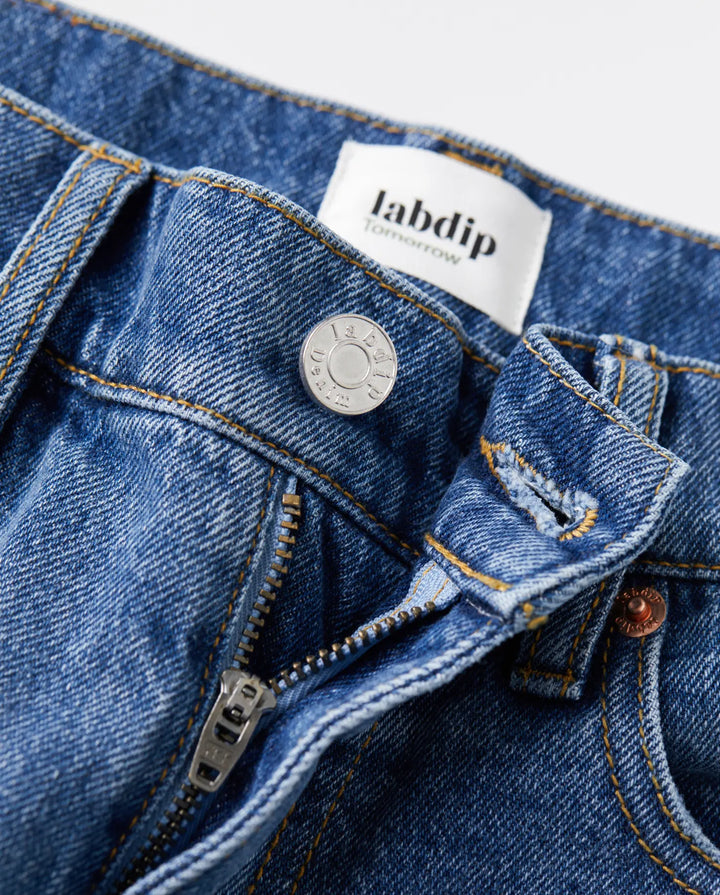 Gros plan sur un jean en denim bleu indigo avec fermeture éclair métallique et bouton argenté, coupe droite adaptée à toutes les morphologies féminines.