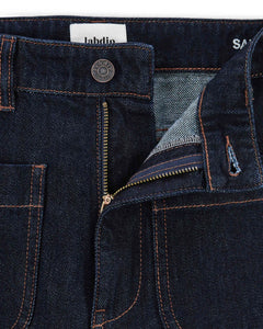 Jean femme en denim brut foncé 100% coton avec une coupe droite et un tissu épais et robuste, fermeture éclair métal et coutures apparentes couleur cuivrée.