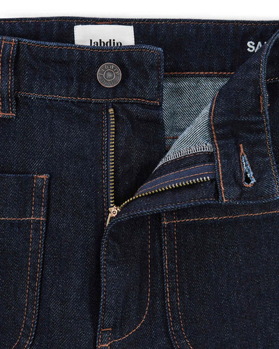 Jean femme en denim brut foncé 100% coton avec une coupe droite et un tissu épais et robuste, fermeture éclair métal et coutures apparentes couleur cuivrée.