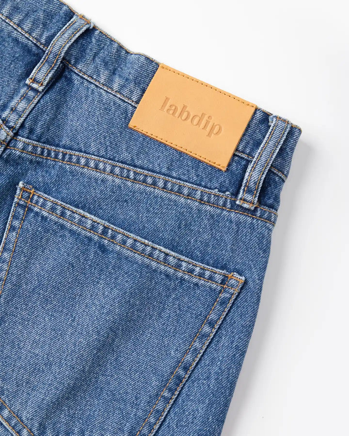 Jean en denim bleu clair 100% coton avec coupe droite et taille haute, finitions soignées et étiquette en cuir beige discrète.