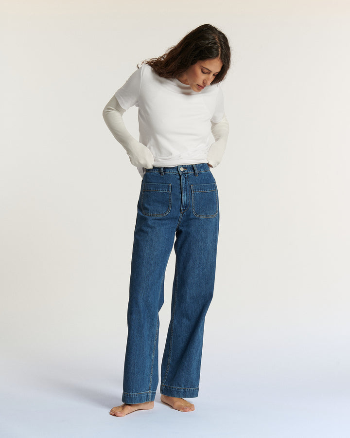 Femme debout portant un jean droit en denim bleu 100% coton avec poches plaquées, combiné à un haut blanc simple, dans un décor minimaliste lumineux.
