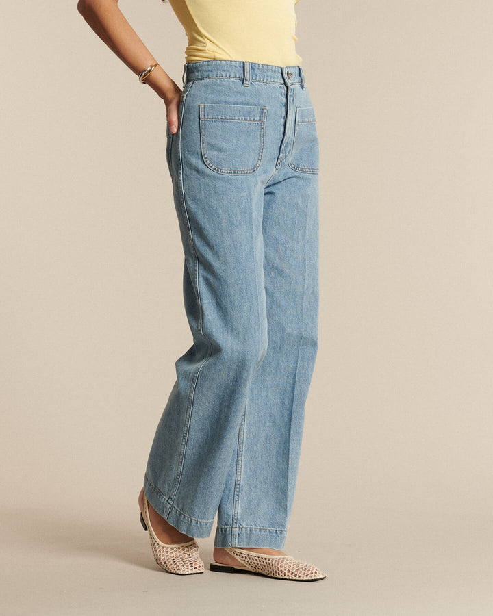 Femme portant un jean droit en denim bleu clair 100% coton, avec poches plaquées à l'avant, associé à un haut jaune et des chaussures ouvertes beiges.